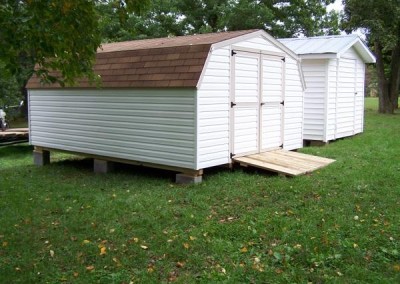 A white, vinyl shed with a shingled, barn style roof. Shed has a wooden ramp and a set of solid double doors.