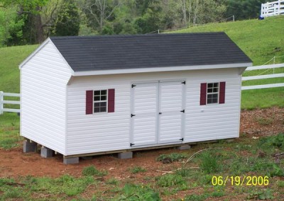 A white vinyl shed with white trim and a carriage style shingled roof. Shed has two windows with shutters and a set of double doors.