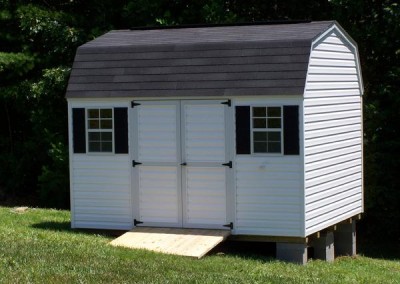 A 10x12 white vinyl shed with a shingled, barn style roof. Shed has a set of double doors and two windows with a treated wooden ramp