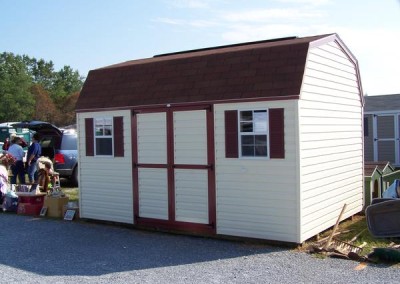 A white vinyl shed with a shingled, barn style roof. Shed has two windows with shutters and solid, vinyl double door