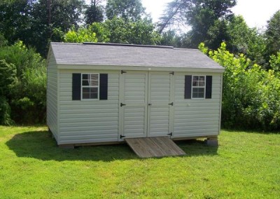 A vinyl, 10x16 shed with an a-roof style, shingle roof. Shed has double door, two windows, and a wooden ramp