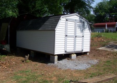A white vinyl mini barn with a barn style, shingled roof. Shed has no windows and a set of double doors.