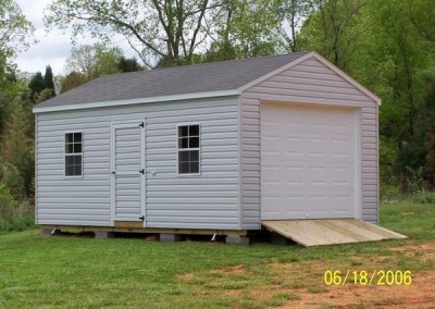 A 12x12 vinyl shed with a shingled roof. Shed has a garage door and wooden ramp, the shed has a 3 foot wide door and two windows.