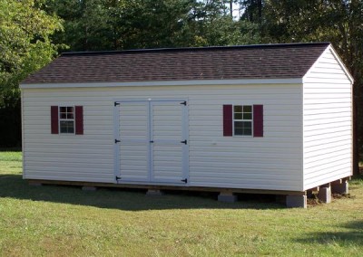 A vinyl 12x12 shed with an a-roof style shingled roof. Shed has a solid double vinyl door and two windows with maroon shutters.