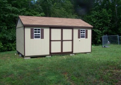 A painted shed with a shingled, a-roof style roof. Shed has brown trim, two windows with shutters, and a solid double door