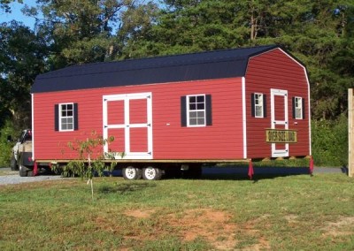 A red vinyl, 14x28 size shed with a black metal, barn style roof. Shed has two large windows and a set of double doors along one side. On gable end, the shed has a 3 foot vinyl door and two smaller windows on either side. Shed is loaded on a trailer for transport.