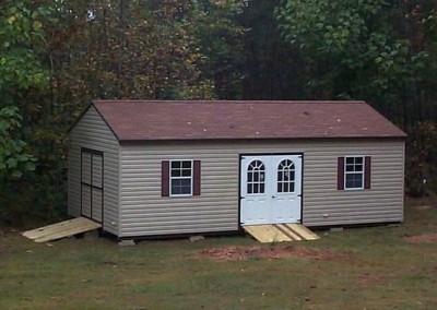 A 14x28 sized vinyl shed with an a-roof style roof. Shed has a double door on gable end with a treated wooden ramp. Shed side has two windows on either side of a double circle top house door with a treated wooden ramp