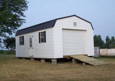 A white vinyl shed with a barn style, metal roof. Shed has a circle top house dor and two windows with shutters. Shed has gable vent, a garage door, and a wooden ramp