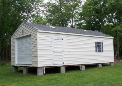 A 14x28 white shed with a 12x12 gable vent and a shingled a-roof style roof. The roof has two skylights. The shed has two windows with shutters and a 3 foot door with a garage door