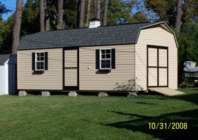 A vinyl high barn with a shingled, barn style roof. Shed has a 3 foot wide vinyl door and two windows on side. A set of double doors with a wooden ramp are on the gable end. Shed has a cupola on roof