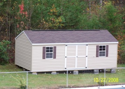 A vinyl shed with a a-roof style shingled roof. Shed has double doors, two windows with shutters and a ridge vent