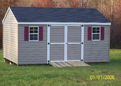 A vinyl shed with white trim and an a-roof style, shingled roof. Shed has maroon shutters and a treated wooden ramp