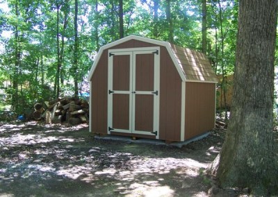 A brown painted mini barn with a shingled, barn style roof. Shed has no windows and a set of double doors. Shed has white trim