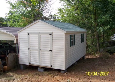 A vinyl shed with 2 windows and a set of double doors. The roof is an a-roof style and is shingled. The shed has two windows and two shutters.