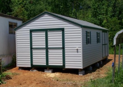 A 12x24 Vinyl shed with green trim and an a-roof style shingled roof. Shed has a double door on gable end and a two windows and a double door on side