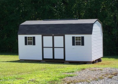 A 10x20 white vinyl shed with a shingled, barn style roof. Shed has a ridge vent, a solid vinyl double door and two windows with shutters