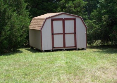 A painted mini barn with a shingled, barn style roof. Shed has no windows and a set of double doors