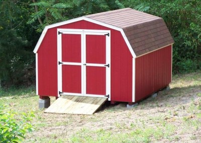 A red painted mini barn with a shingled, barn style roof. The shed has no windows, but has a set of double doors and a treated wooden ramp