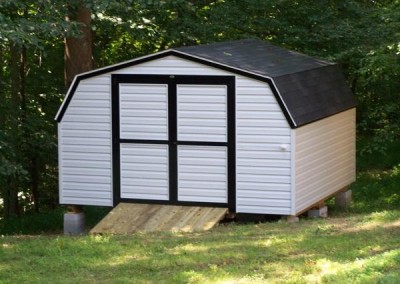 A white mini barn with bak trim. Shed has a solid double door and ramp and a barn style, shingled roof
