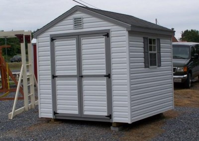 An 8x8 white vinyl shed, with a double door and two windows with shutters and a 8x8 white gable vent. Shed has a shingled a-roof style roof