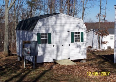 A white vinyl shed with a set of 4 foot wide double doors. Shed has two windows and a wooden ramp. The roof is a barn style and is shingled.