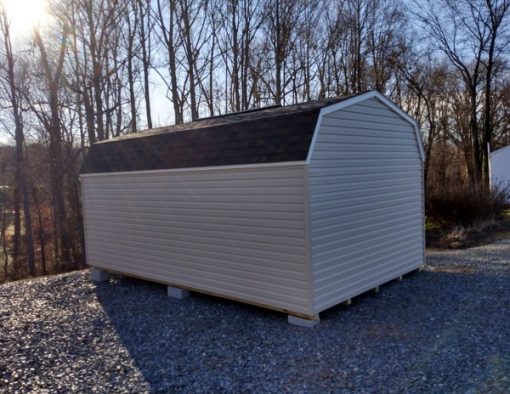 12x20 size vinyl high barn roof style shed with tan siding, white trim, brownwood architectural shingle roof, brown shutters. Has 6 foot fiber solid shed doors and two windows.