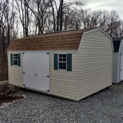 12x16 size vinyl high barn roof style shed with almond siding, white trim, desert tan architectural shingle roof, green shutters. Has 6 foot fiber solid shed doors and two windows.