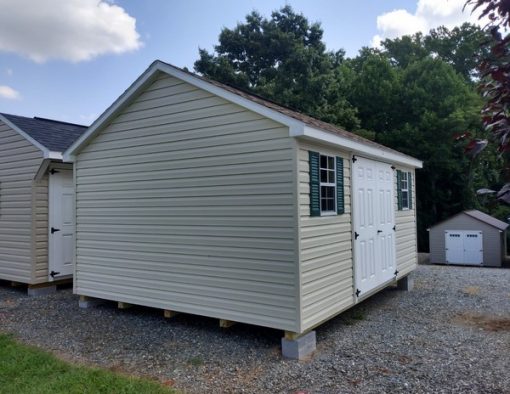12x16 size vinyl classic roof style shed with almond siding, white trim, desert tan architectural shingle roof, green shutters. Has ridgevent, 6 foot fiber doors and two windows.