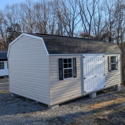 12x20 size vinyl high barn roof style shed with tan siding, white trim, black architectural shingle roof, black shutters. has (2) 8' ridge vents, fiber solid 6 foot double doors and two windows.