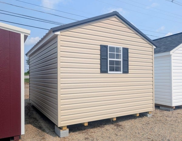 10x12 size vinyl a-roof style shed with tan siding, white trim, black metal roof, black shutters. has 6 foot fiber double doors and two windows.