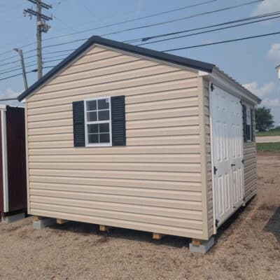 10x12 size vinyl a-roof style shed with tan siding, white trim, black metal roof, black shutters. has 6 foot fiber double doors and two windows.