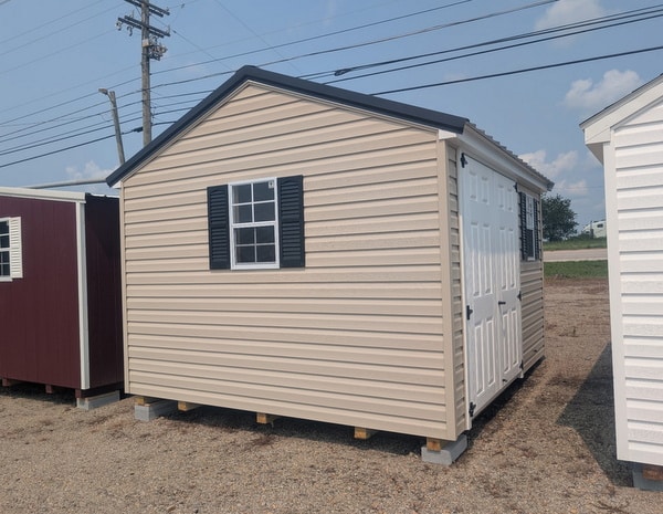 10x12 size vinyl a-roof style shed with tan siding, white trim, black metal roof, black shutters. has 6 foot fiber double doors and two windows.
